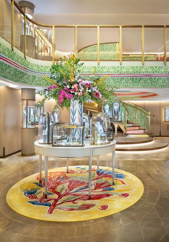 Luxurious boutique interior with vibrant floral centerpiece, elegant staircase, and glass display cases on a colorful rug.