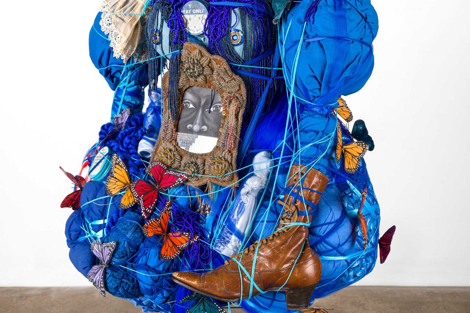 Colorful abstract sculpture with blue fabric, butterflies, a brown shoe, a framed face image, and various intertwined objects.