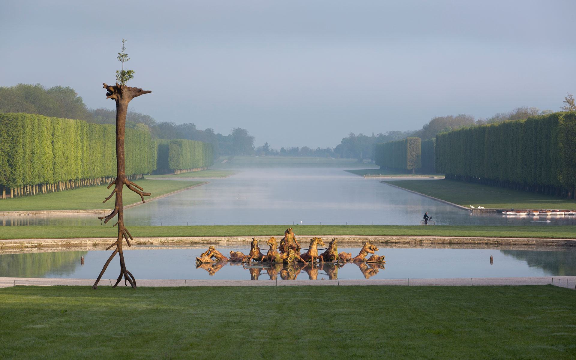 View of formal gardens with a central reflecting pool lined by neatly trimmed trees and a unique tree sculpture in the foreground.