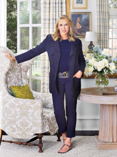 Person standing confidently in a stylish blue outfit, leaning against a patterned armchair in a well-decorated room.