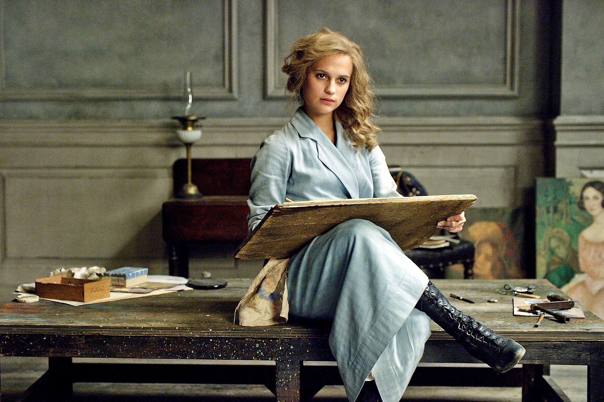 Artist in a blue dress seated at a wooden table, surrounded by art supplies, holding a drawing board in a studio setting.