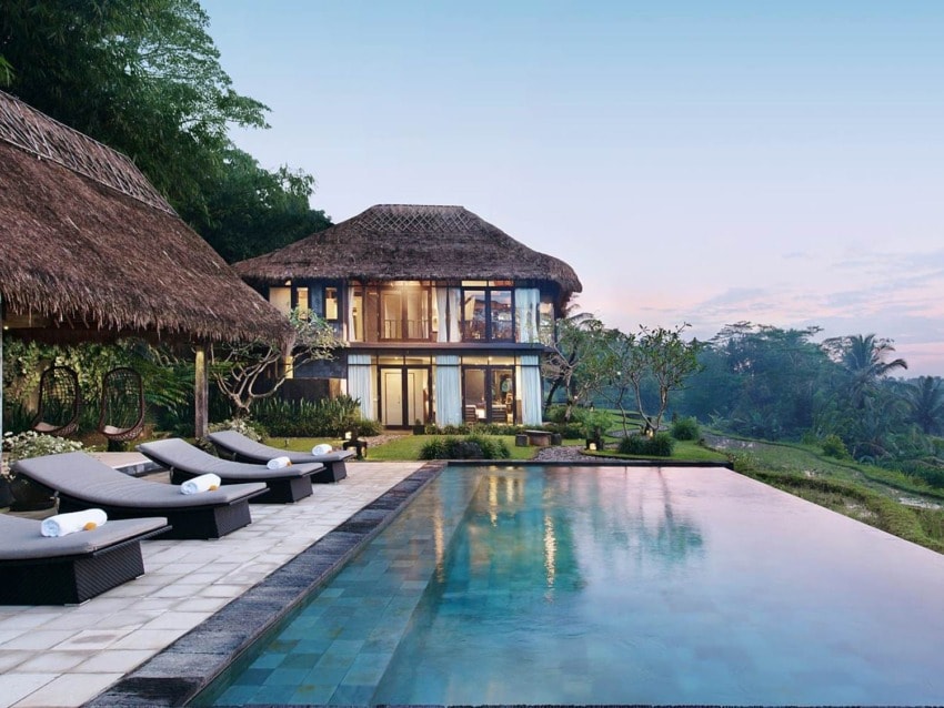 Luxury villa with thatched roof, modern poolside loungers, and a scenic view of lush greenery at sunset.
