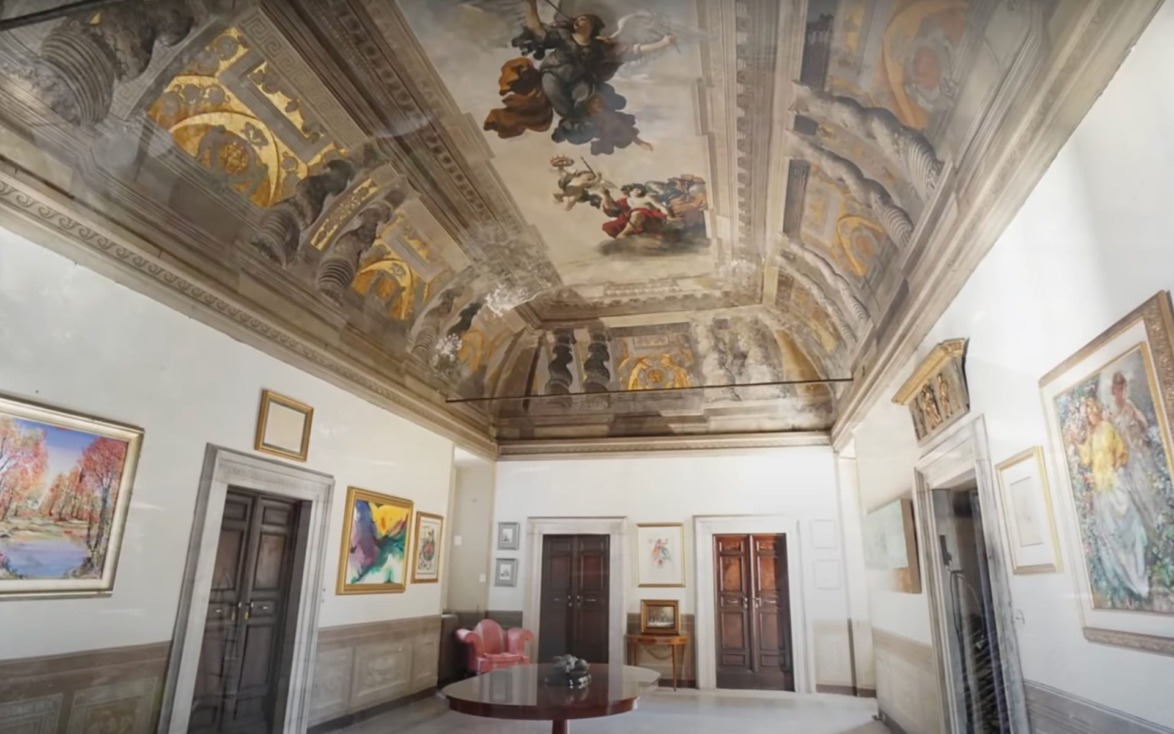 Ornate room with frescoes on a high ceiling, paintings on walls, wooden doors, and a round table with a red chair.