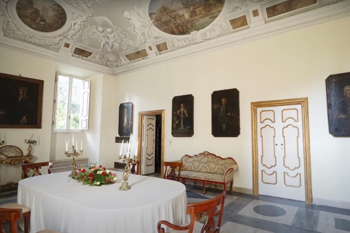 Elegantly decorated historical room with ornate ceiling, classical paintings, and a table with a floral centerpiece.