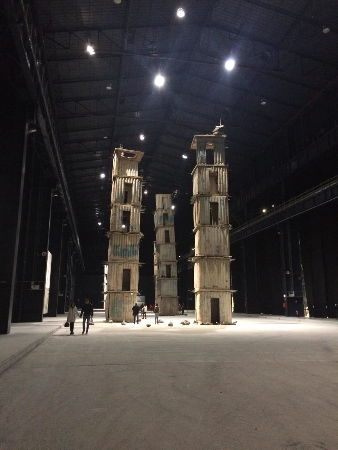 Tall wooden structure installations in a dimly lit spacious gallery, people nearby for scale.