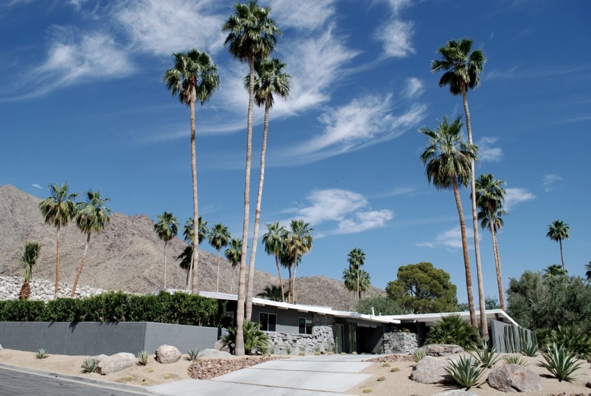 Mid-century modern house with palm trees against a clear blue sky and mountains in the background.