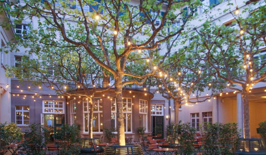 Outdoor courtyard with string lights hanging from trees, surrounded by buildings with windows and outdoor seating.