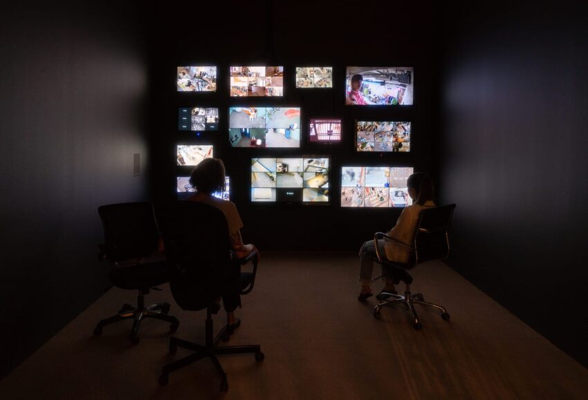 Two people sit in a dark room watching multiple security monitors displaying various surveillance footage.