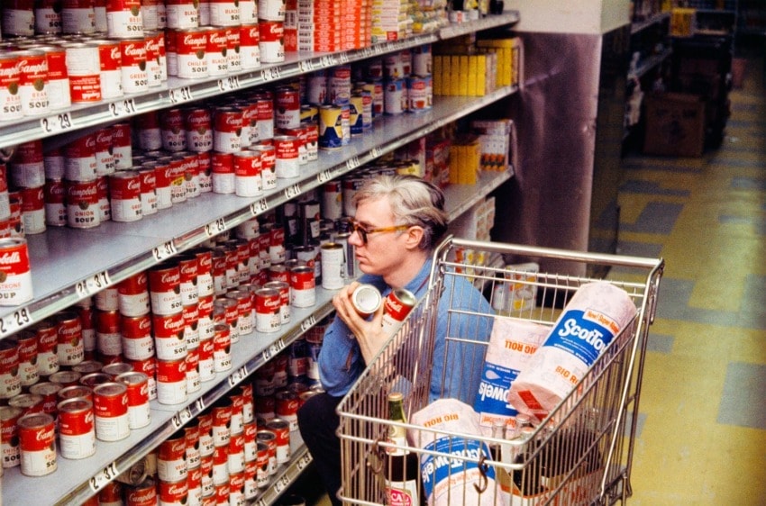 Person in blue shirt shopping for cans in a grocery store aisle, with a shopping cart containing paper towels.