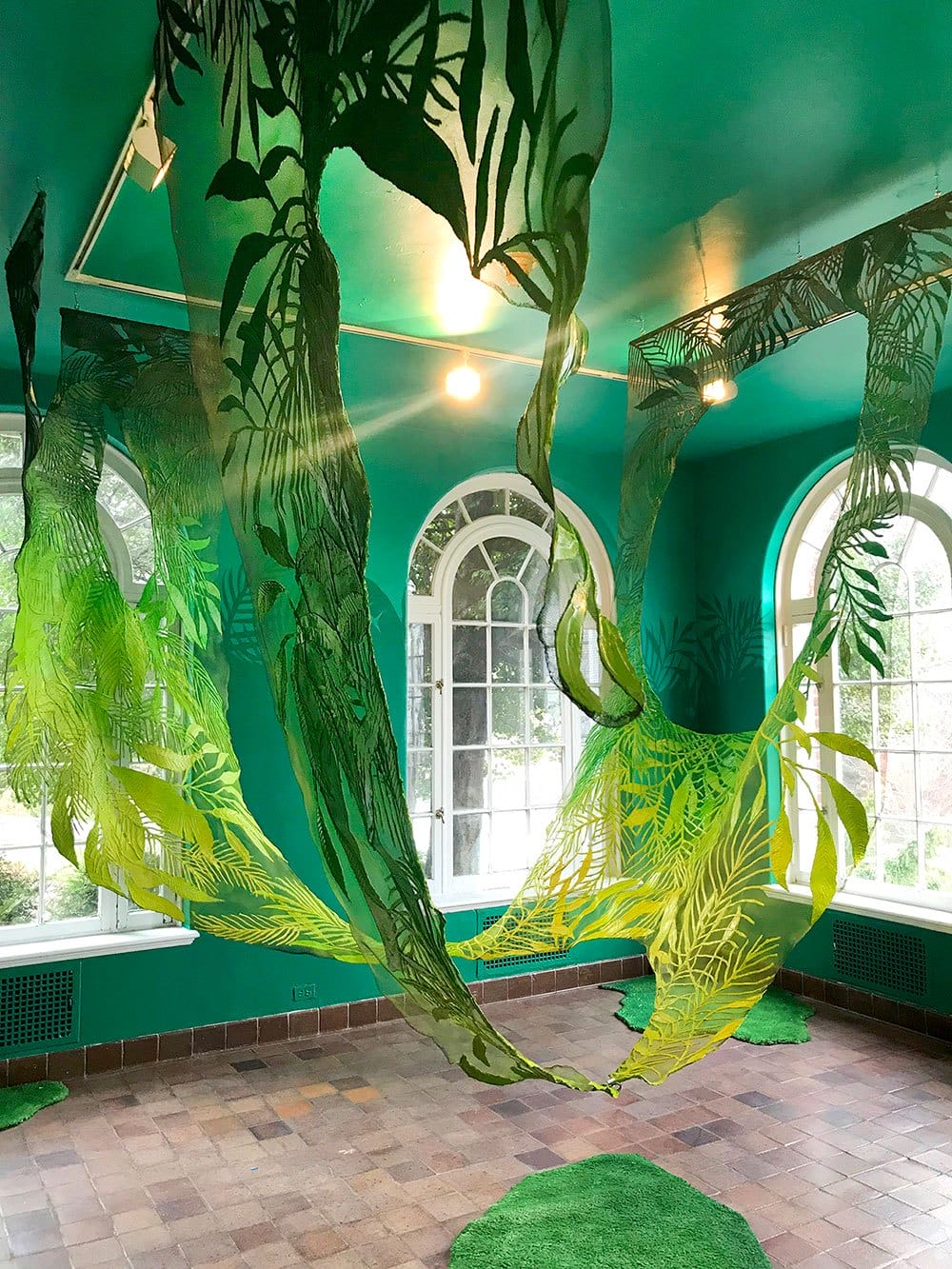 Green art installation with hanging plant motifs in a room with arched windows and brick floor.