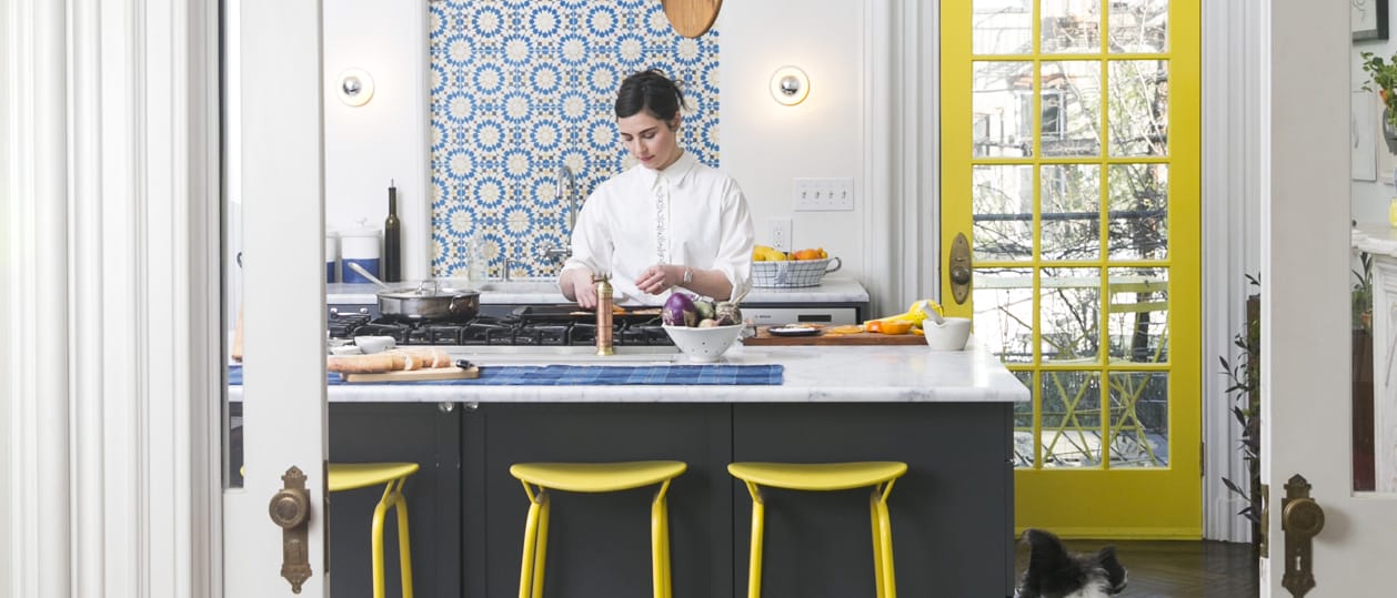 Chef preparing food in a modern kitchen with yellow stools, blue tile backsplash, and bright yellow door in the background.