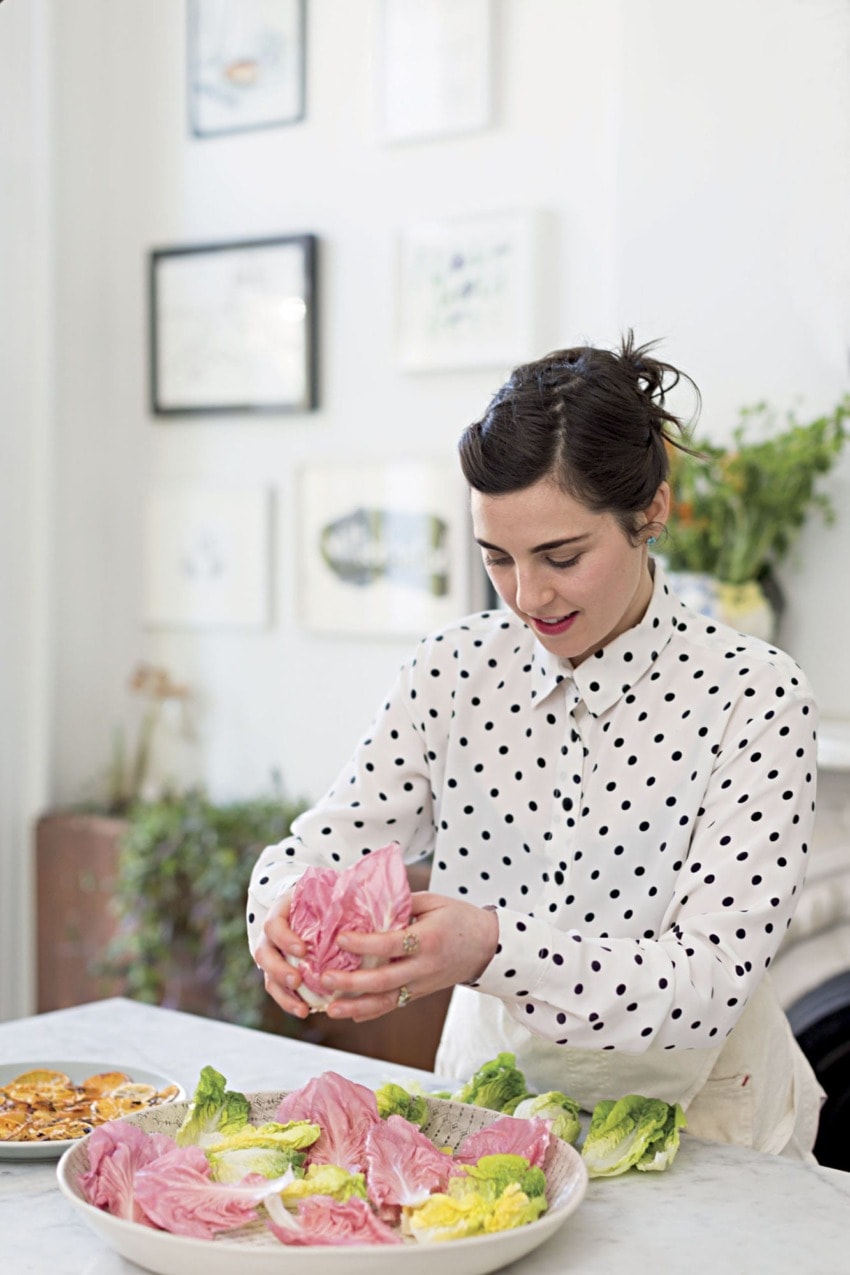 Woman arranging pink and green radicchio leaves in a bowl on a kitchen counter with decorative wall art in the background.