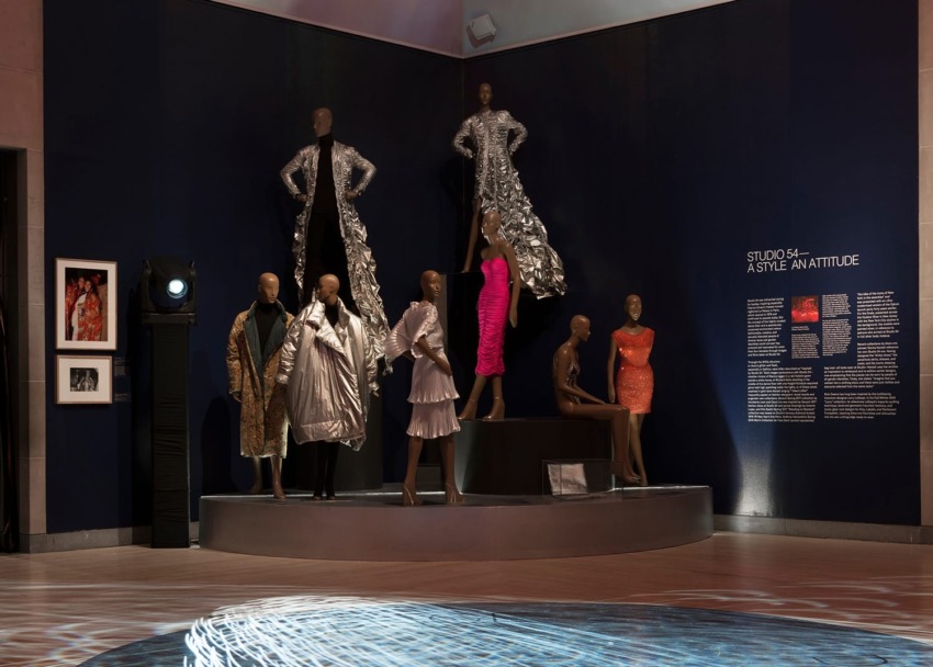 Exhibit featuring Studio 54 fashion with mannequins dressed in glamorous outfits under dramatic lighting.