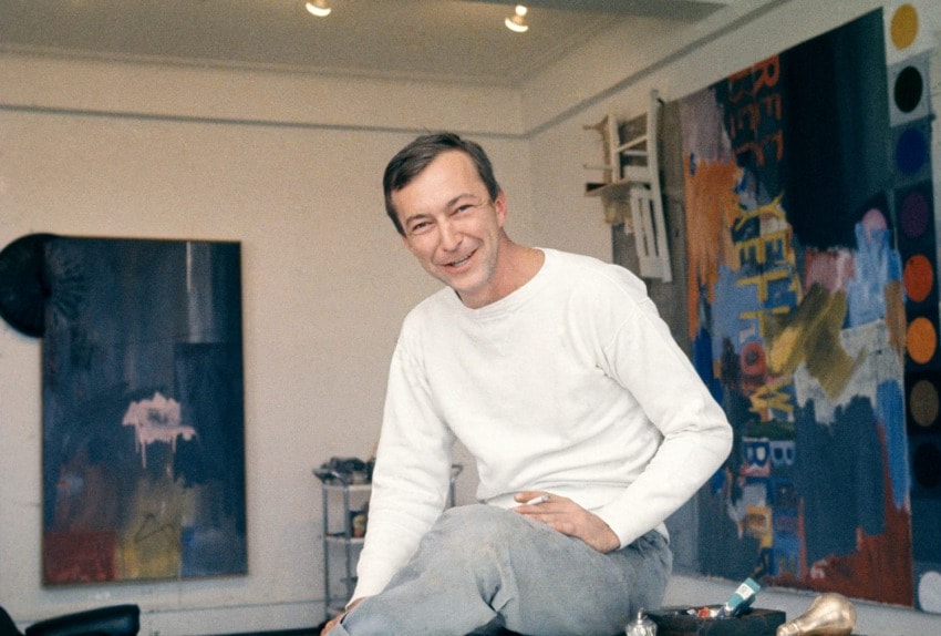Artist seated in a studio with colorful abstract paintings in the background, wearing a white shirt and holding a cigarette.