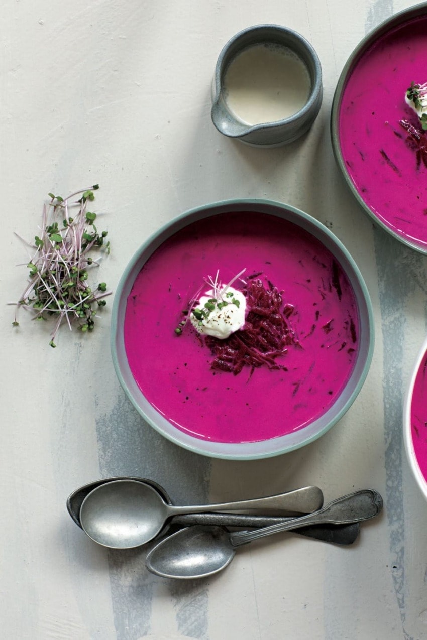 Vibrant pink beet soup in a bowl garnished with cream and herbs, accompanied by a small jug and silver spoons.