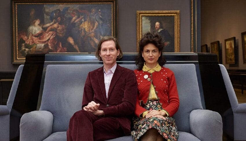 Two people sitting on a couch in an art gallery with framed paintings on the walls in the background.