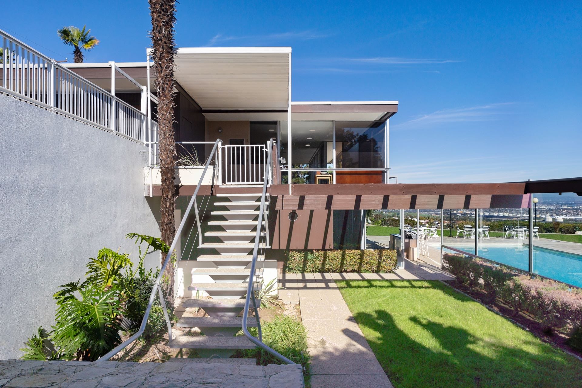 Modern house with staircase, large windows, and a swimming pool in sunny weather, surrounded by greenery and clear blue sky.