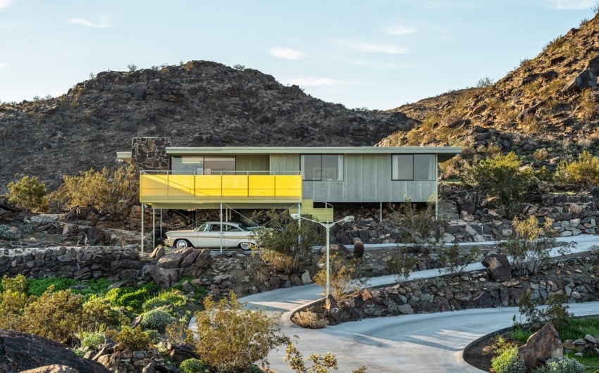 Mid-century modern house on rocky hillside with vintage car and desert scenery under clear sky.
