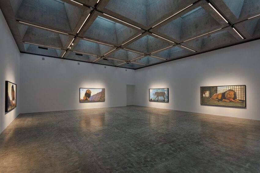 Art gallery interior with framed animal paintings on concrete walls and a high ceiling illuminated by soft, ambient lighting.