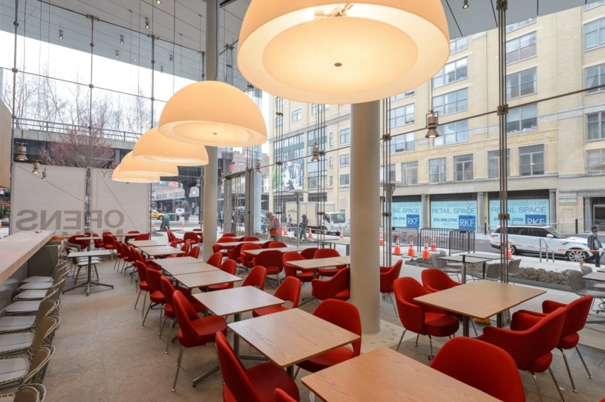 Modern café interior with large windows, red chairs, and wooden tables, overlooking a city street with buildings.