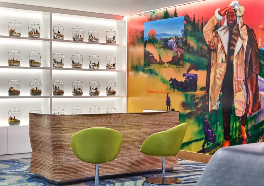 Modern room with two green chairs, a wooden desk, colorful wall art, and a display of jars with small figurines on shelves.