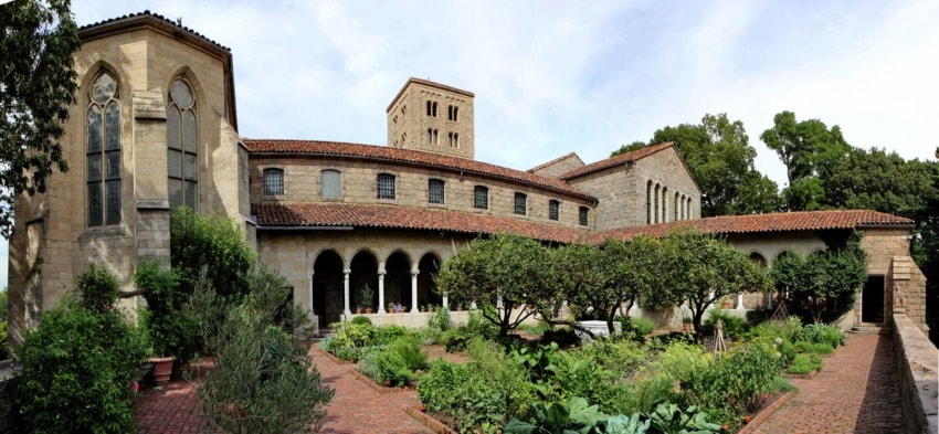 Exterior view of a historic stone building with arched windows, surrounded by lush greenery and a brick pathway in a courtyard.