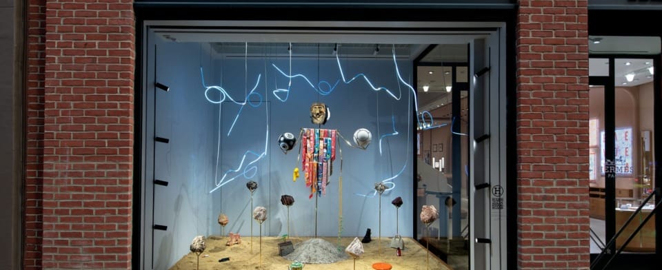 Store display with abstract art installation, featuring hanging objects and neon lights in a street-facing window frame.