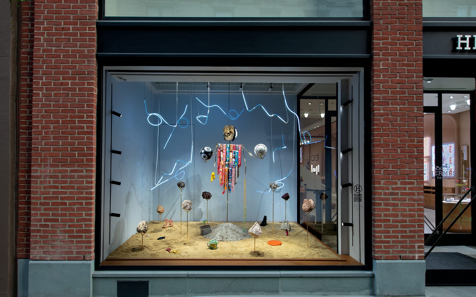 Store display with abstract art installation, featuring hanging objects and neon lights in a street-facing window frame.