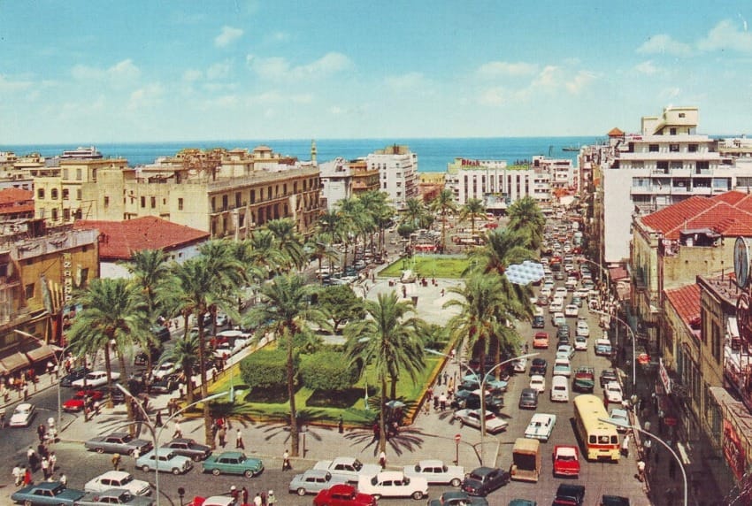 Aerial view of a busy city square with palm trees, surrounded by buildings and traffic, near a coastline under a clear blue sky.