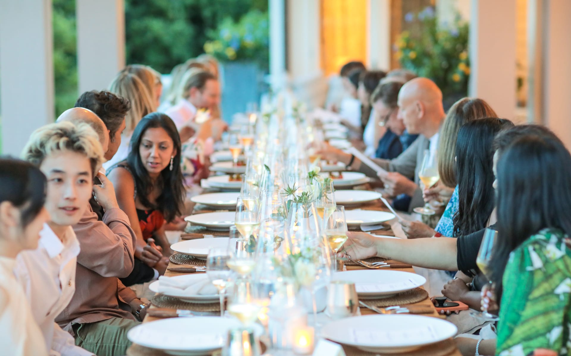 People seated at a long outdoor table set for a meal with decorative plates and glasses, engaging in conversation.