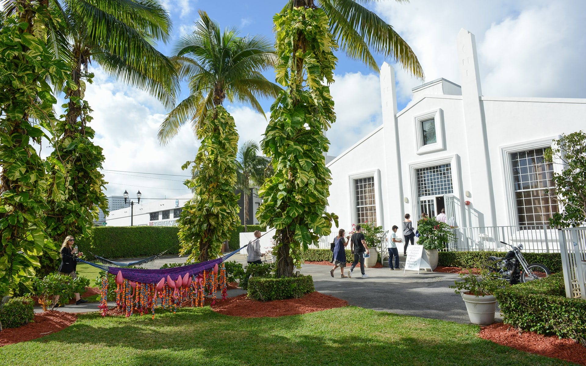 Tropical setting with palm trees and people walking towards a white building with a hammock in the foreground.