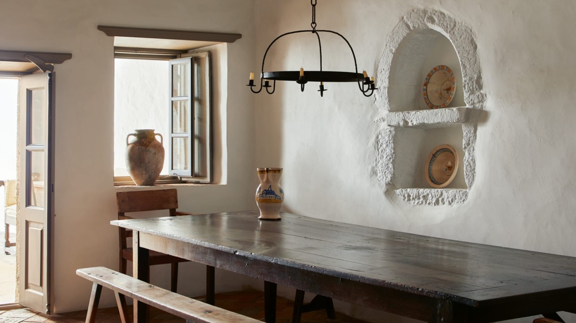 Rustic dining room with a wooden table, benches, pottery, and a vintage chandelier next to an open window and door.