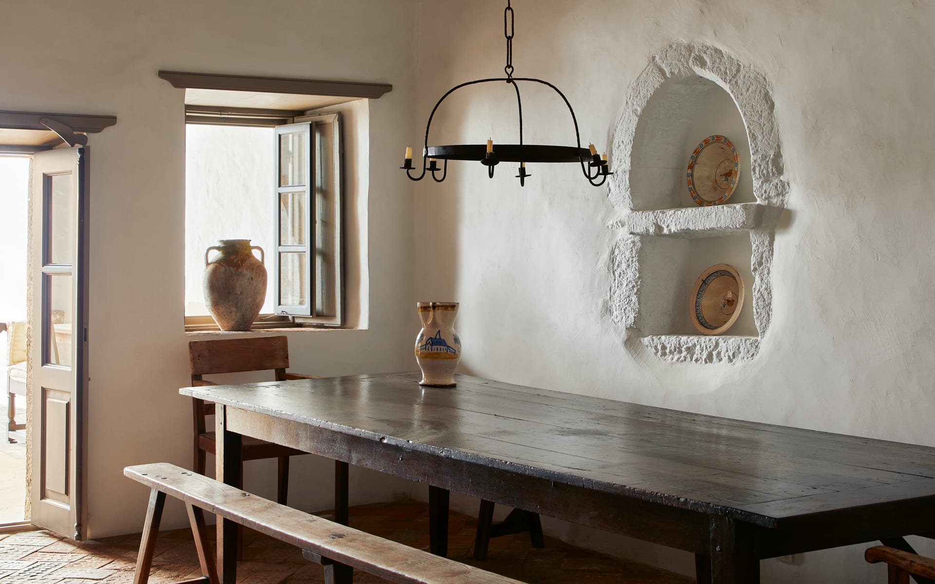 Rustic dining room with a wooden table, benches, pottery, and a vintage chandelier next to an open window and door.