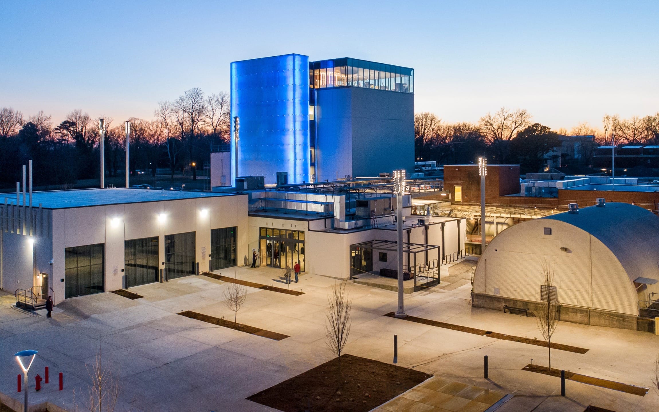 Modern industrial facility at dusk with blue illuminated tower and surrounding trees.