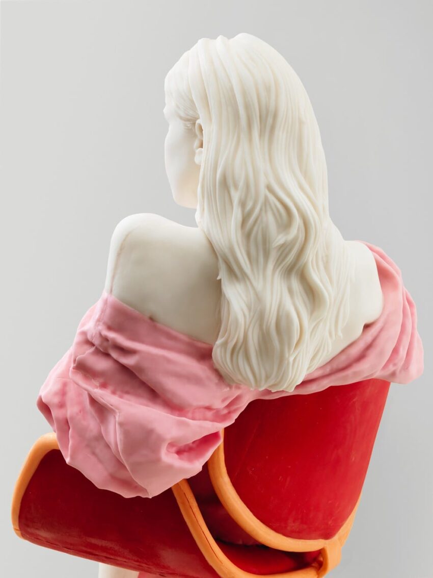 Sculpture of a woman with long hair, draped in a pink fabric, seated on a red chair, viewed from behind.