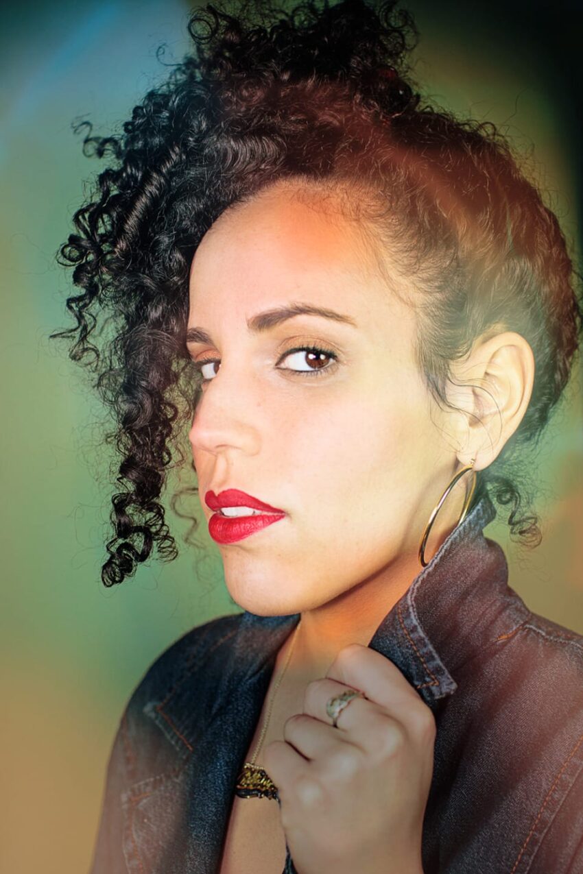 Woman with curly hair and red lipstick, wearing a denim jacket and hoop earrings, looking to the side.