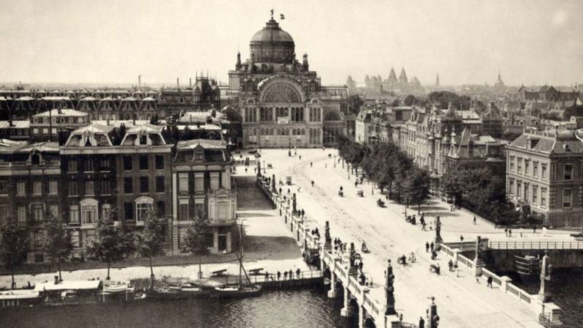 Historic black and white photo of Amsterdam's Paleis voor Volksvlijt and surrounding buildings along a canal bridge.