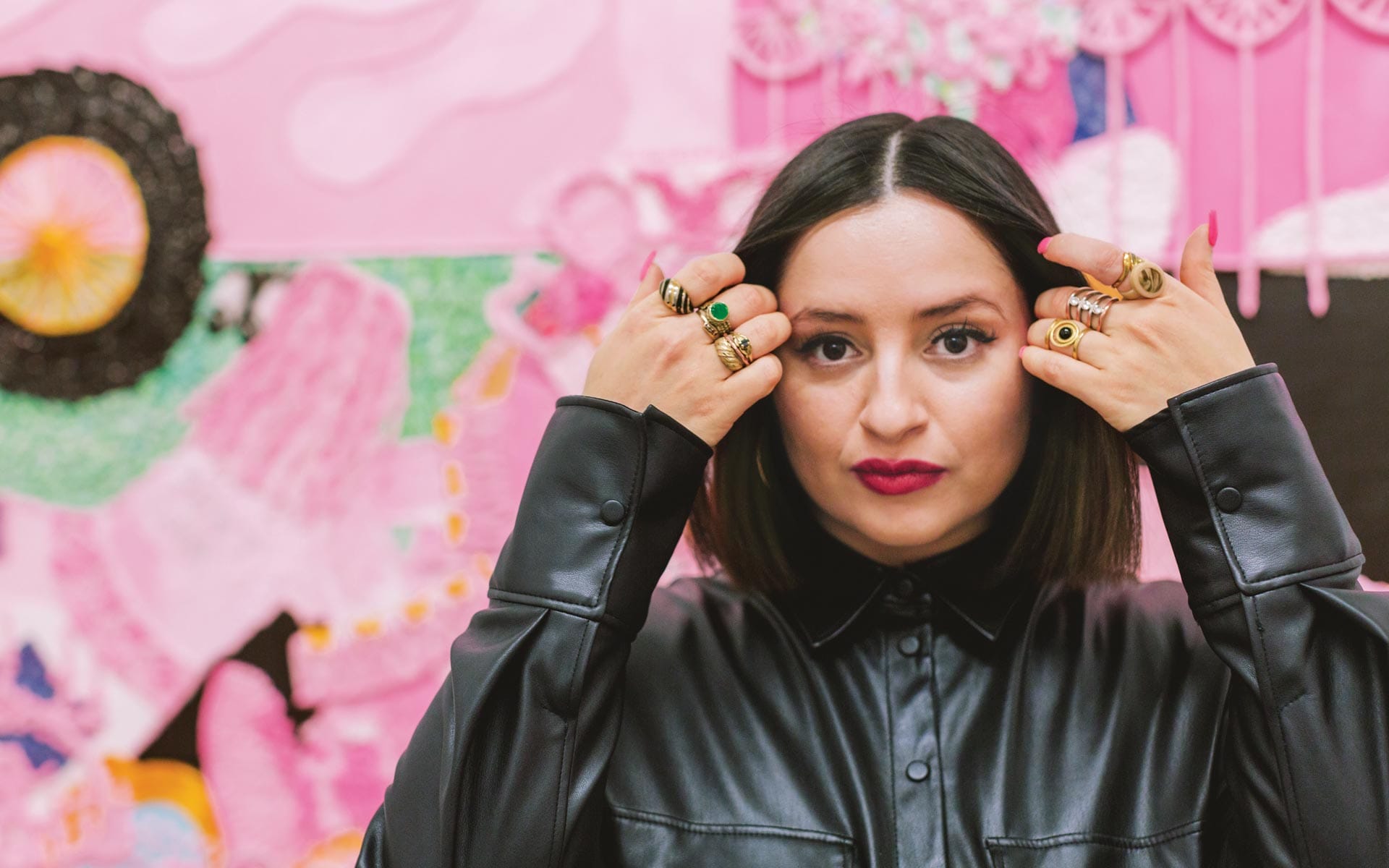 Woman in a black leather shirt with rings, standing in front of a colorful abstract wall art.