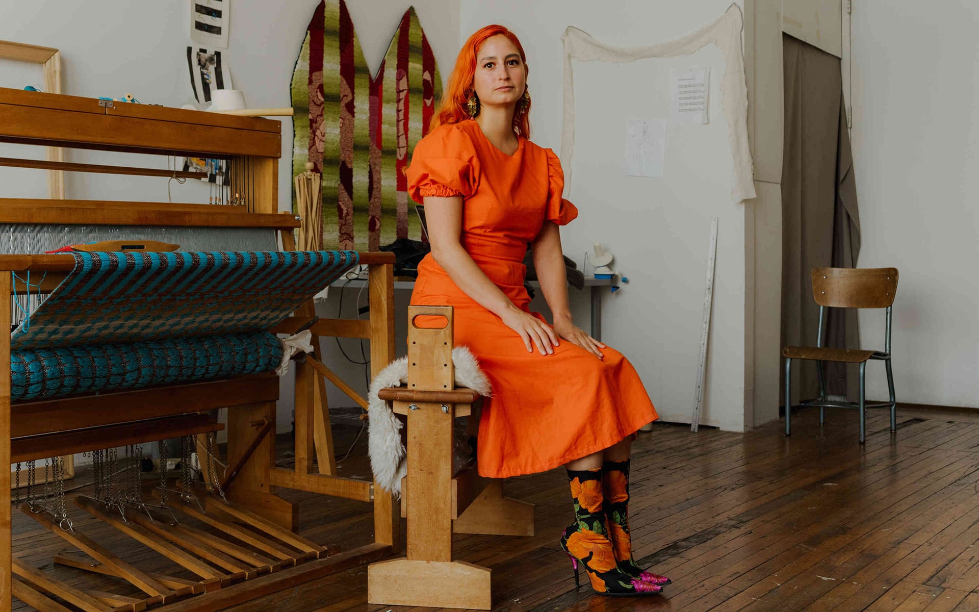 Person in an orange dress sitting in an art studio with weaving equipment and colorful fabric in the background.