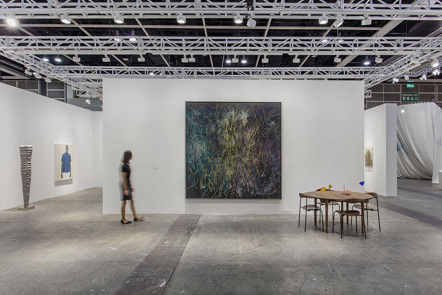 Modern art gallery with abstract painting on display, woman walking by, sculptures and table with chairs in a spacious setting.