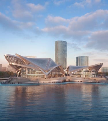 Futuristic waterfront building with curved design, surrounded by water and three glass skyscrapers in the background under a blue sky.