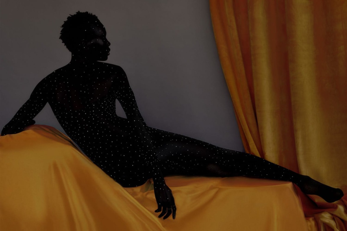 Silhouetted figure covered in sparkling dots reclines on a yellow fabric, with golden curtains in the background.