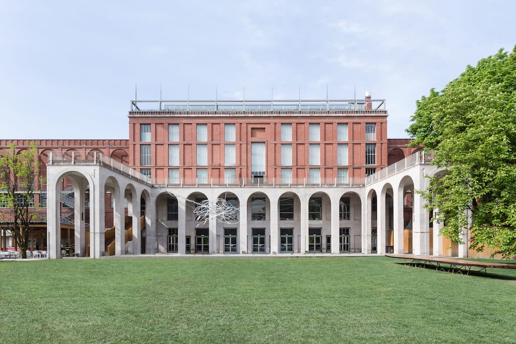 Red brick building with large arched windows, set in a green lawn, and framed by trees under a clear sky.