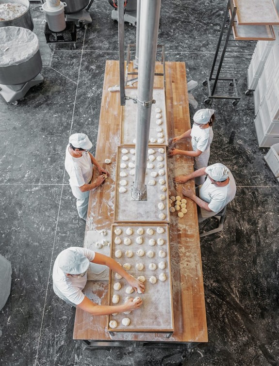 Workers in a bakery forming dough balls on a long wooden table viewed from above.