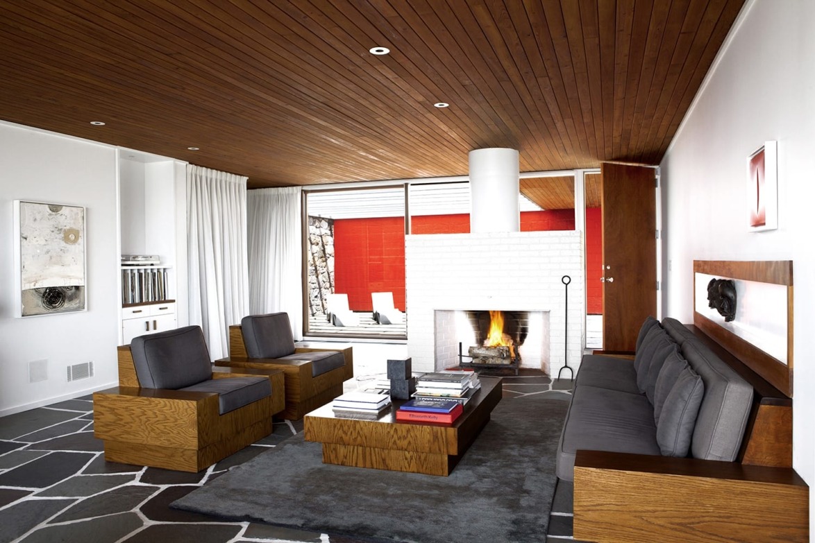 Modern living room with wooden ceiling, gray furniture, stone floor, and a lit brick fireplace, offering a cozy atmosphere.