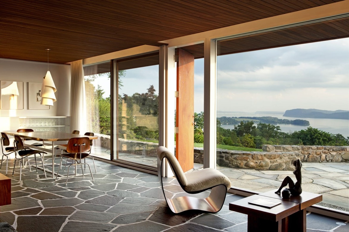 Modern living room with stone floor, stylish furniture, large glass windows, and scenic view of a lake and mountains.