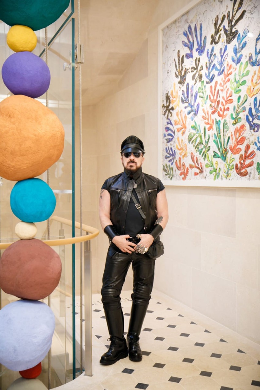 Person in black leather outfit standing in front of a colorful artwork on a tiled floor with round sculptures nearby.