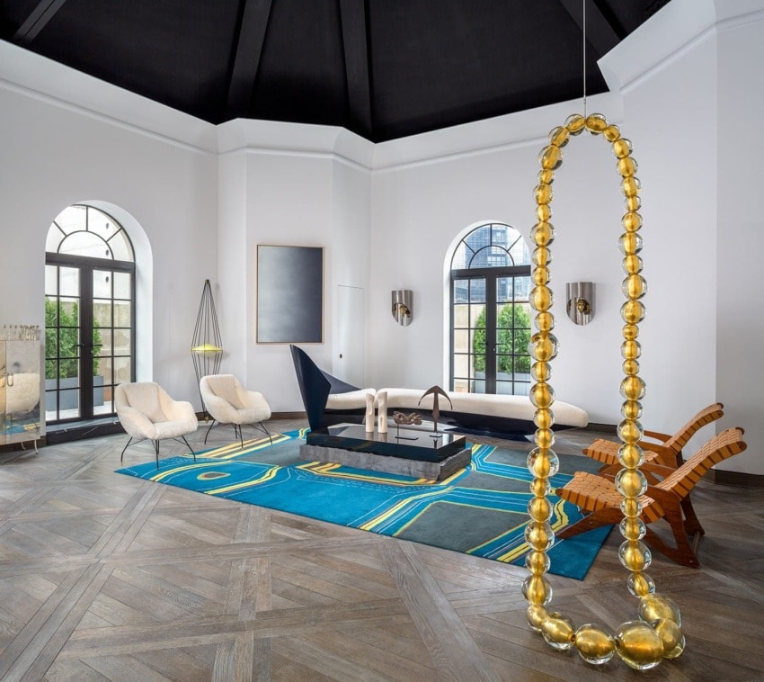 Modern living room with blue rug, unique gold sculpture, wooden chairs, and contemporary decor elements.