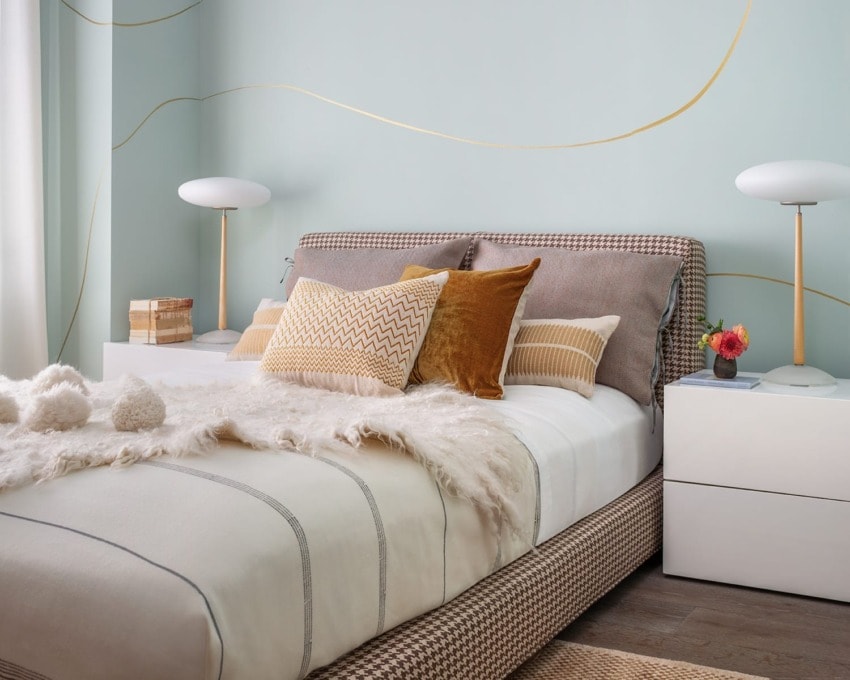 Stylish bedroom with a cozy bed, decorative pillows, fluffy throw, modern lamps, and a light blue wall with gold accents.