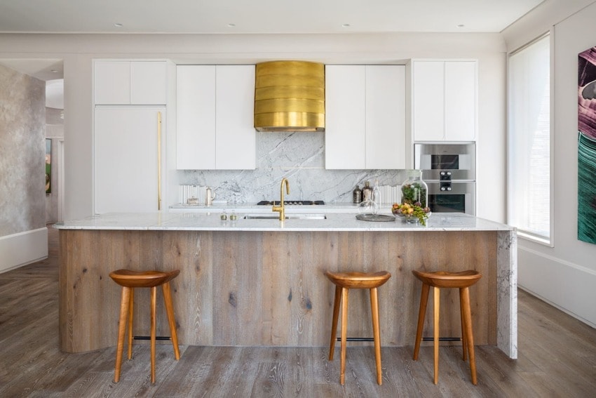 Modern kitchen with wooden island, marble countertop, gold fixtures, and white cabinets. Three wooden stools in front.